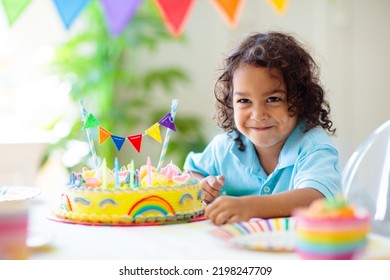 Kids Birthday Party. Children Celebrate With Colorful Cake And Gifts. Little Curly Boy Blowing Candles And Opening Birthday Presents. Friends Play With Rainbow Confetti. Party Home Decoration.