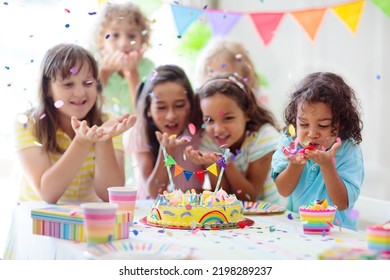 Kids Birthday Party. Children Celebrate With Colorful Cake And Gifts. Little Curly Boy Blowing Candles And Opening Birthday Presents. Friends Play With Rainbow Confetti. Party Home Decoration.