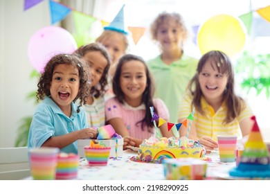 Kids Birthday Party. Children Celebrate With Colorful Cake And Gifts. Little Curly Boy Blowing Candles And Opening Birthday Presents. Friends Play With Rainbow Confetti. Party Home Decoration.