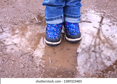 Kids Boots In Muddy Puddle. Colorful Winter Outdoor Rain Shoes On Childrens Feet In Action. A Child On A Walk Stay In The Mud. Choosing Footwear For An Active Lifestyle. Autumn Rainy Weather.