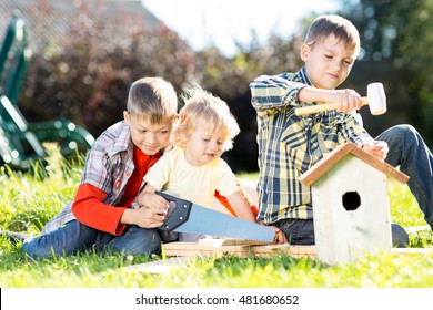 Kids Boys Making Birdhouse Together Sitting On The Grass. Oldest Child Teaches Youngest Brother To Work With Tools