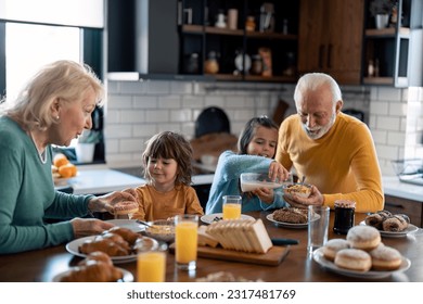 Kids at breakfast with grandparents. Little girl adding milk into cereal bowl with grandpas help while grandma is serving donut on a plate to her little brother. Cheerful family moments at home. - Powered by Shutterstock