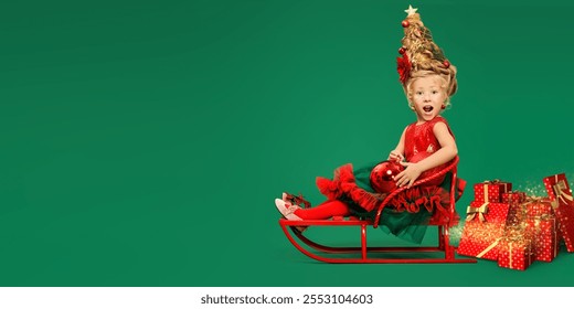 Kid's Christmas and New Year. Funny emotional little girl in a festive dress and xmas tree hairstyle rides a sled with many gifts. Green background with copy space. 