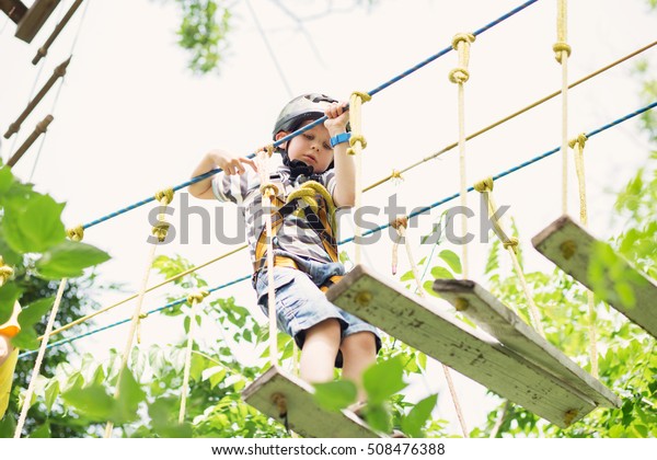 Kids climbing in adventure park. Boy enjoys\
climbing in the ropes course adventure. Child climbing high wire\
park. Happy boys playing at adventure park, holding ropes and\
climbing wooden stairs.