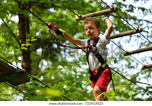 Kids climbing in adventure park. Boy enjoys\
climbing in the ropes course adventure. Child climbing high wire\
park. Happy boys playing at adventure park holding ropes and\
climbing wooden stairs