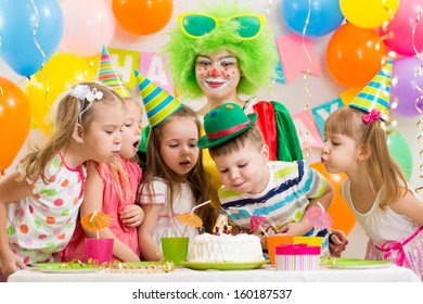 Kids With Clown Celebrating Birthday Party And Blowing Candle On Cake