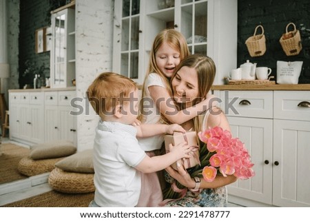 Kids congratulates mother and gives present, bouquet of tulip flowers in kitchen at home. Mother's day concept. Mom hugging children and surprised. Greeting card. International Women's Day. Back view