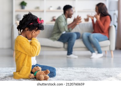 Kids And Domestic Conflicts. Little African American Girl Crying While Her Parents Arguing At Home, Upset Black Female Child Sitting On Floor With Teddy Bear And Covering Face, Selective Focus