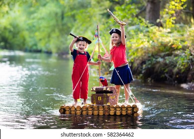 Kids dressed in pirate costumes and hats with treasure chest, spyglasses, and swords playing on wooden raft sailing in a river on hot summer day. Pirates role game for children. Water fun for family - Powered by Shutterstock