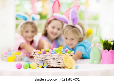 Kids Dyeing Easter Eggs. Children In Bunny Ears Dye Colorful Egg For Easter Hunt. Home Decoration With Flowers, Basket And Rabbit For Spring Holiday Celebration. Little Boy And Girl Decorate Home.