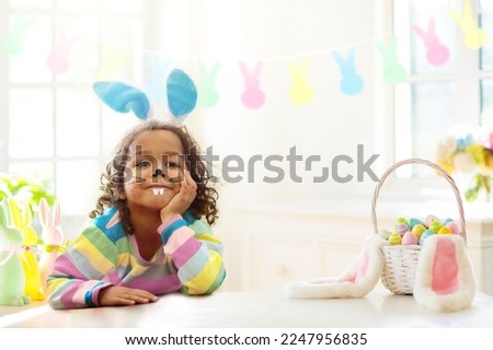 Kids dyeing Easter eggs. Children in bunny ears dye colorful egg for Easter hunt. Home decoration with flowers, basket and rabbit for spring holiday celebration. Little curly boy decorating home.