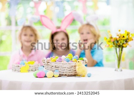 Kids dyeing Easter eggs. Children in bunny ears dye colorful egg for Easter hunt. Home decoration with flowers, basket and rabbit for spring holiday celebration. Little boy and girl decorate home.