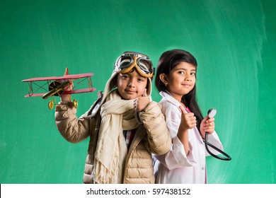 Kids And Education Concept - Small Indian Boy And Girl Posing In Front Of Green Chalk Board In Pilot Fancy Dress And Doctor Costume With Stethoscope, Standing Over Green Chalkboard Background