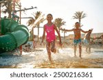 Kids enjoy a fun-filled day at a water park, running and playing in the water splashes. The bright sun, palm trees, and colorful slides create a perfect summer adventure atmosphere.
