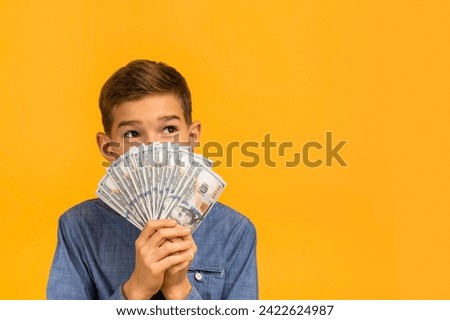 Kids and financial literacy. Rich teen boy holding bunch of cash dollar banknotes, pensive mle kid covering face with money, standing isolated on yellow studio background, copy space