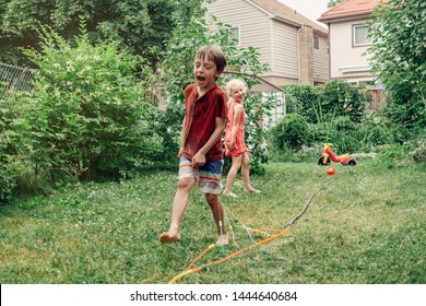 Kids Friends Boy Girl Splashing With Gardening Hose Sprinkler  On Backyard On Summer Day. Children Playing With Water Outside At Home Yard. Candid Authentic Real Life Moment Of Funny Activity