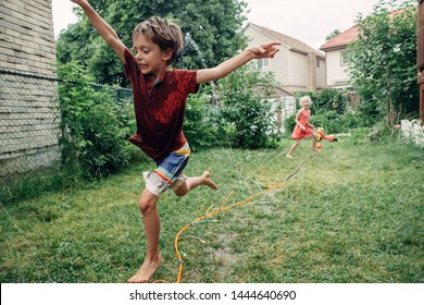 Kids Friends Boy Girl Splashing With Gardening Hose Sprinkler On Backyard On Summer Day. Children Playing With Water Outside At Home Yard. Candid Authentic Real Life Moment Of Funny Family Activity