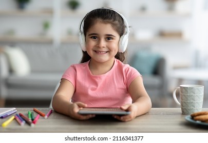 Kids And Gadgets. Portrait Of Cute Preteen Girl In Wireless Headphones Using Digital Tablet While Sitting At Desk At Home, Little Arab Female Child Holding Tab Computer And Smiling At Camera