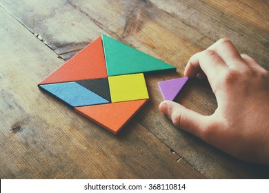 kid's hand holding a missing piece in a square tangram puzzle, over wooden table. 