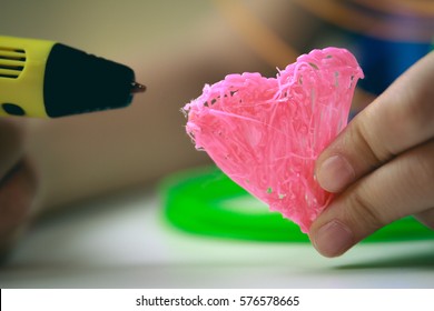 Kids Hand Holding Yellow 3D Printing Pen With Filaments And Makes Heart On White Background. Top View. Copy Space For Text. Selective Focus