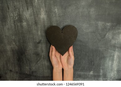 kids hands holding a black wooden heart in a middle of empty chalkboard background