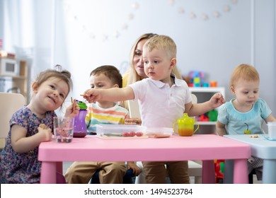 Kids Have A Dinner In Kindergarten. Little Toddlers Boys And Girls From The Group Of Children Sitting At The Table With Lunch And Eat Appetizing. Children With Caregiver In Day Care Centre