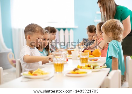 Kids have a dinner in kindergarten. Little boys and girls from the group of children sitting at table with lunch and eat appetizing. Children with caregiver in day care centre. Childs have healthy