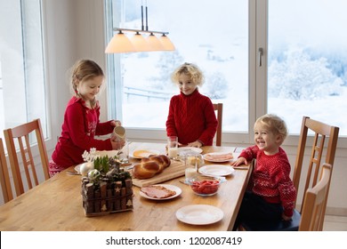 Kids Having Breakfast On Christmas Morning. Family Eating Bread And Drinking Milk At Home On Snowy Winter Day. Children Eat In Sunny Dining Room At Window With Swiss Mountains And Snow View.