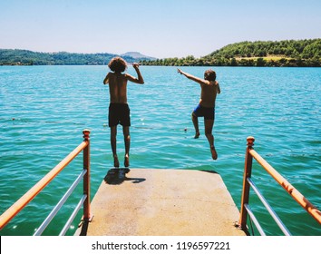 Kids Jumping Off The Dock Into A Beautiful Mountain Lake. Having Fun On A Summer Vacation At The Lake With Friends