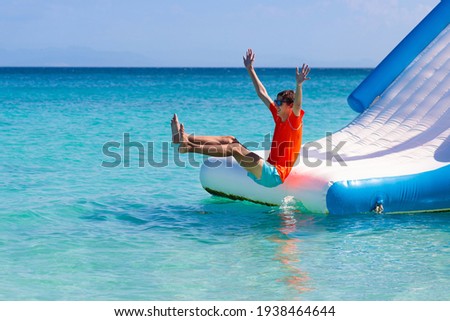 Kids jumping on trampoline on tropical sea beach. Children jump on inflatable water slide. Aqua amusement park in exotic island resort. Family vacation and travel with child. Ocean coast fun.
