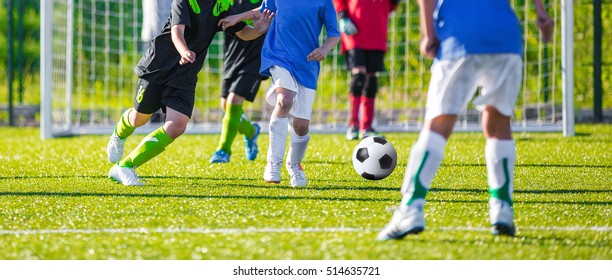 Kids Kicking Football Ball On Sports Field. Young Boys Competing During European Soccer Tournament. Footballers In Blue And Black Shirts.