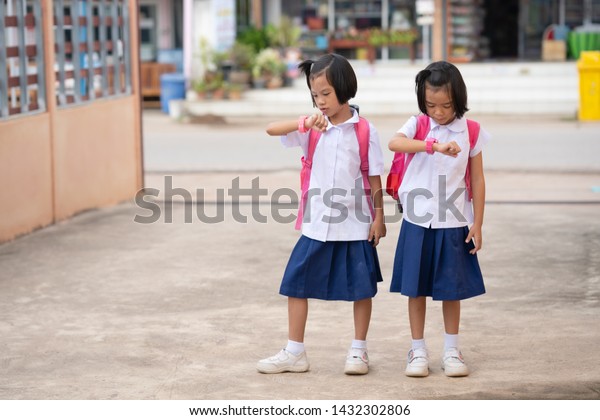 Kids learning concept, kid girls looking wacth on\
school uniform for go to school in morning, they wearing pink\
backpack and pink wacth and white shoes, they standing on home or\
house for waiting car
