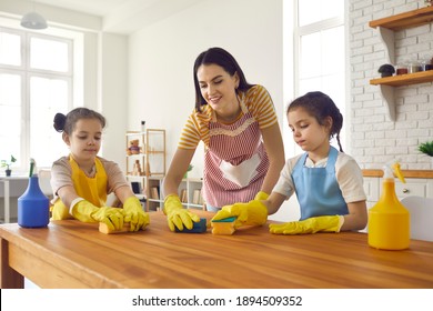 Kids Learning To Do Family Chores. Cute Daughters Helping Mom Wash Wooden Table At Home. Happy Mother And Children Wearing Yellow Rubber Gloves Cleaning Kitchen Surface With Sponges And Detergent