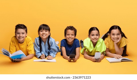 Kids lifestyle and leisure, happy childhood concept. Cheerful multiracial diverse preteen children lying on floor isolated on yellow background, reading books, doing homework, using phones, banner - Powered by Shutterstock