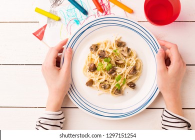 Kid's meal - spaghetti with mushrooms. Colorful dinner - plate in child's hands on white wooden table. Plate captured from above (top view, flat lay).