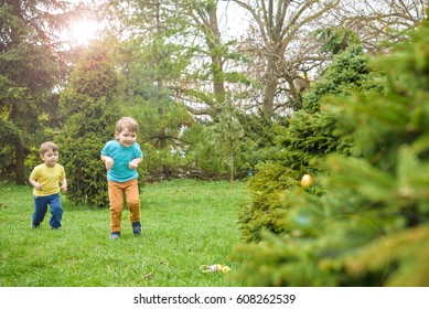 Kids On Easter Egg Hunt In Blooming Spring Garden. Children Searching For Colorful Eggs In Flower Meadow. Toddler Boy And His Brother Friend Kid Boy Play Outdoors.