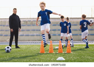 Kids On Sports Soccer Training. Boys Running Balls And Jumping Over Cones. Agility Exercises For Youths In Football Team. Coach Of Football Team Coaching Players. Kids Making Training Drills Program