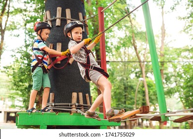 Kids Passing The Cable Route High Among Trees. Young Brothers Climbing In High Rope Course In Adventure Park. Children In Forest Adventure Park. Kids In Safety Helmets, Extreme Sport. Summer Camp.