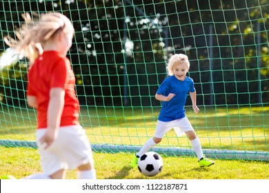 Kids Play Football On Outdoor Field. Children Score A Goal At Soccer Game. Girl And Boy Kicking Ball. Running Child In Team Jersey And Cleats. School Football Club. Sports Training For Young Player.