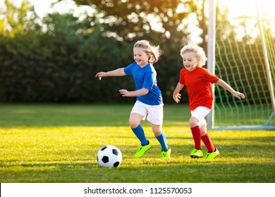 Kids Play Football On Outdoor Field. Children Score A Goal At Soccer Game. Girl And Boy Kicking Ball. Running Child In Team Jersey And Cleats. School Football Club. Sports Training For Young Player.