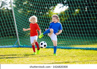 Kids Play Football On Outdoor Field. Children Score A Goal At Soccer Game. Girl And Boy Kicking Ball. Running Child In Team Jersey And Cleats. School Football Club. Sports Training For Young Player.