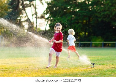 Kids Play With Water On Hot Summer Day. Children With Garden Sprinkler. Outdoor Fun. Boy And Girl Run On Football Field After Training Under Water Drops.