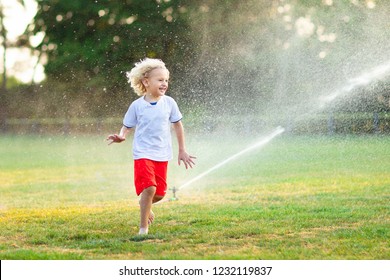 Kids Play With Water On Hot Summer Day. Children With Garden Sprinkler. Outdoor Fun. Boy And Girl Run On Football Field After Training Under Water Drops.