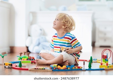 Kids play with wooden railway. Child with toy train. Educational toys for young children. Little boy building railroad tracks on white floor at home or kindergarten. Cute kid playing cars and engine.