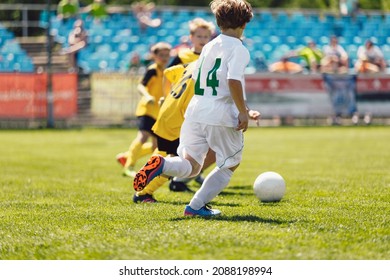 Kids Playing Football In Sunny Day. School Boys Running Fast On Grass Soccer Pitch. Junior Football Players Compete In Tournament Match. Football Spectators, Stadium Crowd In Blurred Background