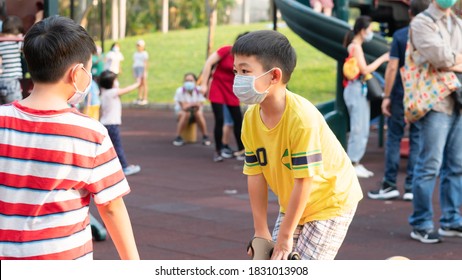 Kids Are Playing Together In The Park During COVID-19 With The Surgical Masks On.Children And Parents At The Playground During Summer Day Time.Outdoor Activity After Lock Down At The Public Play Area.