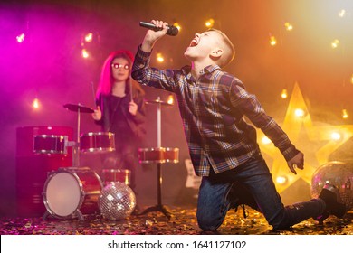 Kids Pretending To Be A Rock Band Play And Sing At Studio Or Stage. Boy Singing And Girl Paying On Drums