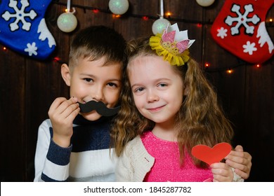 Kids With Props For A Photo Booth. Little Boy And Girl With The Requisite Mustache, Heart And Crown On Christmas Wooden Background. Event, Holiday, Party. Children Congratulate Happy New Year.