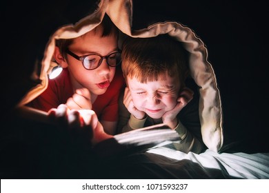 Kids Reading A Book Under The Blanket. The Elder Brother Reads A Scary Tale Aloud. Little Boy In Fright Closed His Eyes And Covered His Ears