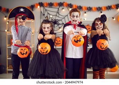 Kids are ready for tricks or treats on Halloween night. Four children standing in room in carnival costumes and spooky make-up stretch their buckets pumpkins to camera. Halloween celebration concept.